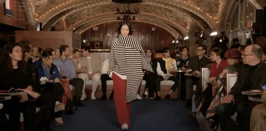 Model walking down runway in red pants and black and white sweater with black and white striped scarf at the Tommy Hilfiger show. 